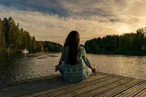 Frau sitzt mit Blick auf den See im Schneidersitz und meditiert